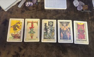 Cartas do deck Rider-Waite representando arquétipos do tarot: O Louco, O Pendurado, A Morte, O Diabo e A Justiça, dispostas sobre mesa escura.